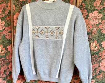 May include: A gray sweatshirt with white stripes and a geometric pattern on the front. The sweatshirt has a crew neck and long sleeves.