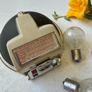 Könnte beinhalten: Vintage Kamera-Belichtungsmesser in Beige und Schwarz mit einer Anzeige der Verschlusszeiteinstellungen für 200er Film. Der Belichtungsmesser hat einen silbernen Metallsockel und wird von zwei Glühbirnen und einer gelben Blume begleitet.