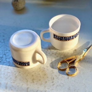 May include: Two white ceramic teacups with blue and gold trim. The cups are sitting on a white surface with a pair of gold scissors.