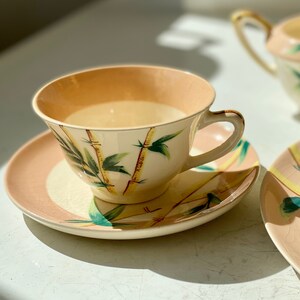 Puede incluir: Un juego de taza y platillo de té vintage con un diseño de bambú. La taza y el platillo son de color crema con un borde rosa y presentan un diseño de bambú verde y amarillo.
