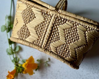 May include: A rectangular woven basket with a handle and a lid. The basket is made of light brown and tan woven material with a geometric pattern. A sprig of greenery and yellow flowers are next to the basket.