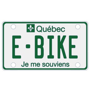 Könnte beinhalten: Ein weißes und grünes Quebec E-Bike-Kennzeichen mit dem Text "Je me souviens" unten.
