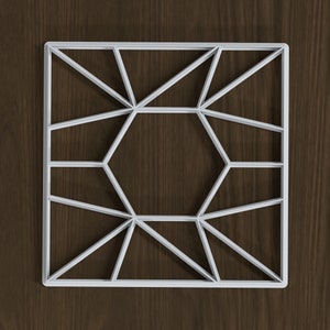 Puede incluir: Un diseño geométrico blanco, con un marco cuadrado con un patrón interior de triángulos y un hexágono central. El diseño se encuentra sobre un fondo de vetas de madera marrón oscuro, creando un contraste.