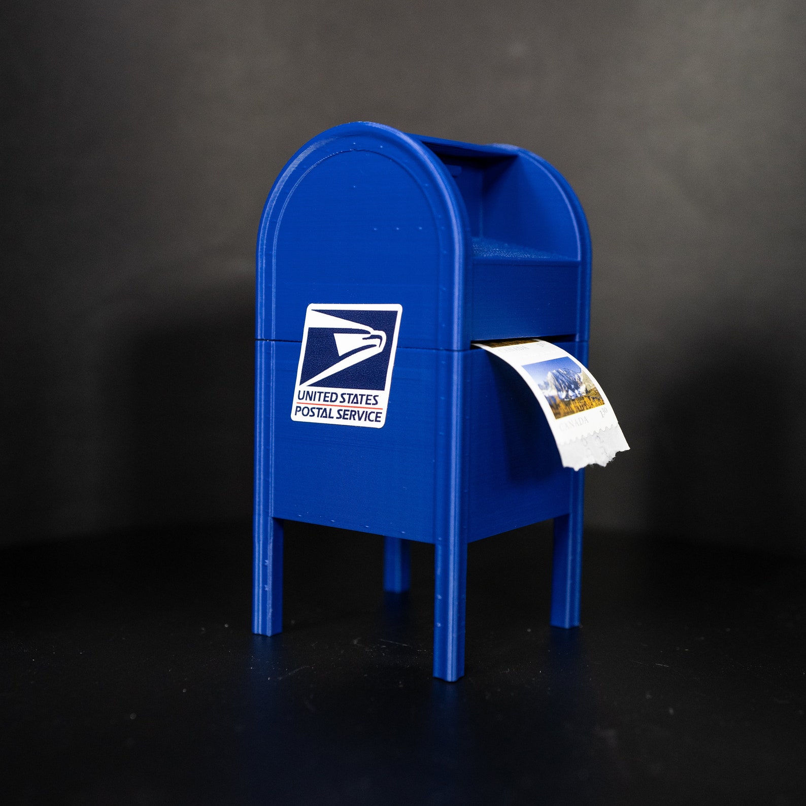 A miniature USPS mailbox stamp dispenser with a stamp peeking out, torn edges