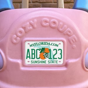 Puede incluir: Coche de juguete de plástico rosa con una matrícula que dice "MYFLORIDA.COM ABC 123 SUNSHINE STATE" y las palabras "Cozy Coupe" en relieve en la parte superior.