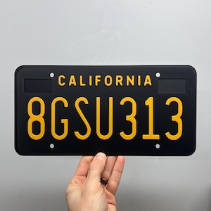 Può includere: Targa nera della California con scritte gialle. La targa riporta "CALIFORNIA" in alto e "8GSU313" al centro. La targa è tenuta da una mano.