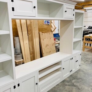 May include: White entertainment unit with open shelves, closed cabinets, and a drawer. The unit has black hardware and is made of wood.