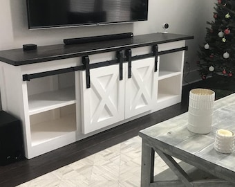 May include: White entertainment center with two sliding barn doors, black metal hardware, and a dark wood top. The doors have a white X-shaped design. The center has two shelves on each side.