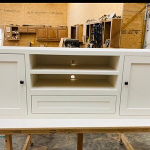 May include: White entertainment unit with two doors, two shelves, and a drawer. The doors and drawer have black knobs. The unit is sitting on a wooden table.