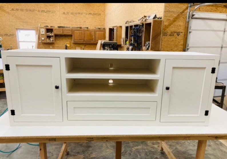May include: White entertainment unit with two doors, two shelves, and a drawer. The doors and drawer have black knobs. The unit is sitting on a wooden table.