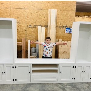 May include: White entertainment unit with three open shelves on each side, a central cabinet with two shelves and a drawer, and three doors with black knobs on each side. The entertainment unit is in a room with wood panelling.
