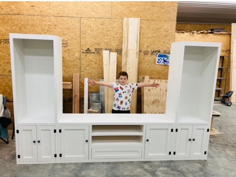 May include: White entertainment unit with three open shelves on each side, a central cabinet with two shelves and a drawer, and three doors with black knobs on each side. The entertainment unit is in a room with wood panelling.