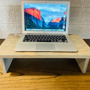 May include: A light wood laptop stand with a laptop on top. The laptop is silver and has a screen displaying a mountain range. The stand is on a wooden surface.