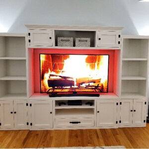 May include: White entertainment unit with a fireplace-style TV stand. The stand has a built-in fireplace with a glowing orange fire and a black TV screen. The entertainment unit has two white cabinets on each side of the TV stand.