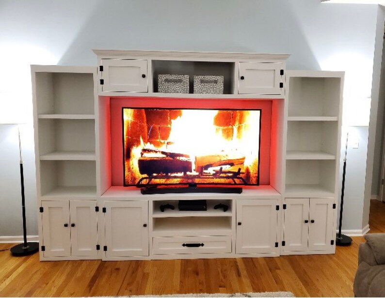 May include: White entertainment unit with a fireplace-style TV stand. The stand has a built-in fireplace with a glowing orange fire and a black TV screen. The entertainment unit has two white cabinets on each side of the TV stand.