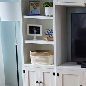 May include: White wooden cabinet with two doors and three shelves. The shelves are decorated with framed photos, a small plant, a woven basket, and a blue geometric sculpture. The cabinet has black hinges and knobs.