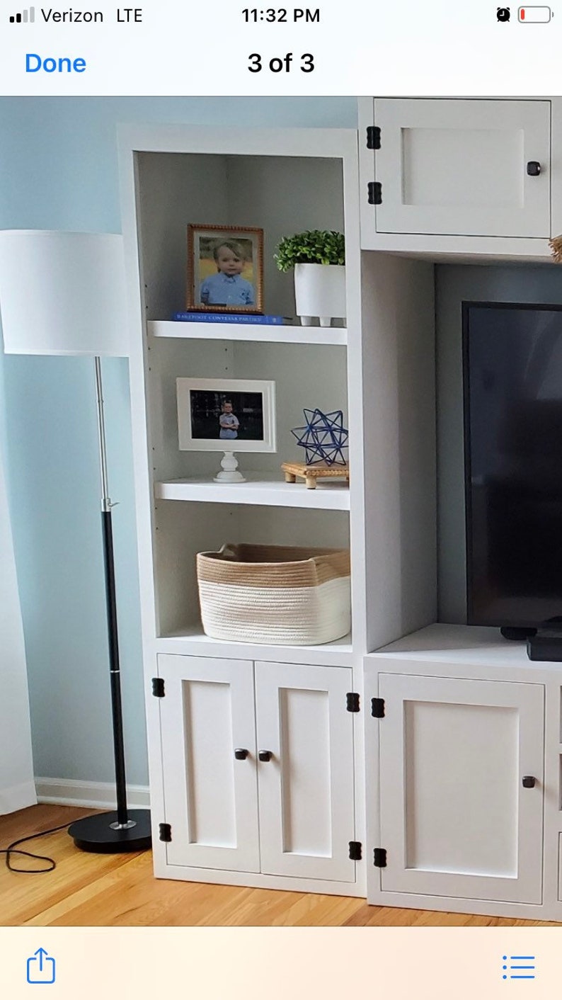 May include: White wooden cabinet with two doors and three shelves. The shelves are decorated with framed photos, a small plant, a woven basket, and a blue geometric sculpture. The cabinet has black hinges and knobs.
