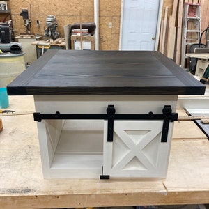 May include: A white wooden coffee table with a black top and a sliding barn door style door. The door has a white X design and black hardware. The table is sitting on a wooden surface.