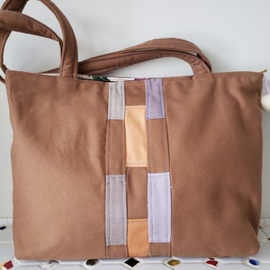 May include: A brown canvas tote bag with a patchwork design of purple, gray, and orange fabric strips. The bag has two brown straps and a zipper closure.