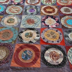Op de afbeelding: Een quilt gemaakt van verschillende stoffen met een patchwork-ontwerp. De quilt is gemaakt van vierkanten met cirkels in het midden. De cirkels zijn gemaakt van verschillende stoffen en hebben verschillende patronen. De quilt is donkerpaars.