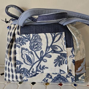 May include: A patchwork shoulder bag with a variety of blue and white fabrics. The bag features a floral print pocket, a striped handle, and a blue and white checkered pattern on the side. The bag is a mix of textures and patterns.