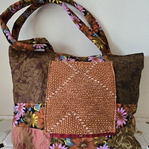 May include: A patchwork tote bag with a brown floral pattern, a central tan panel, and floral accents. The handles are brown with a floral design. The bag features a variety of textures and colors, including pink, black, and gold.