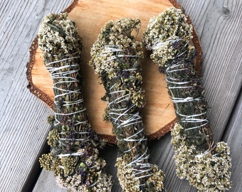Può includere: Tre mazzi di erbe secche legate con spago, disposti su una fetta di legno. I mazzi sono un mix di fiori e foglie secche verdi e bianche.
