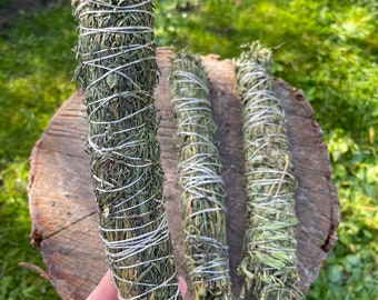 Può includere: Tre mazzi di erbe secche legate con spago, probabilmente per la purificazione o l'incenso. I mazzi sono verdi e marroni e sono su una superficie di legno.