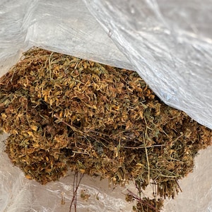 May include: A transparent plastic bag containing dried herbs. The herbs are a mix of brown, yellow, and green, with visible stems and small flowers. The bag is partially open, revealing the contents.