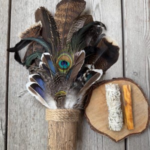 Puede incluir: Un manojo de varitas de incienso con una variedad de plumas, incluidas plumas de pavo real, atadas a un palo de madera. El manojo está al lado de un disco de madera con una varita de incienso de salvia blanca y una varita de palo santo.