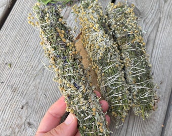 Può includere: Quattro mazzi di erbe secche legate insieme con uno spago. I mazzi sono fatti di erbe verdi e gialle, e alcuni hanno piccoli fiori viola.