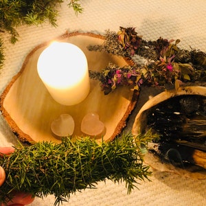 Könnte beinhalten: Ein Holztablett mit einer brennenden weißen Kerze, zwei rosa herzförmigen Kristallen und einem Bündel getrockneter Kräuter. Neben dem Tablett steht eine Keramikschale mit einem schwelenden Bündel getrockneter Kräuter. Im Vordergrund befindet sich ein grüner Zweig.