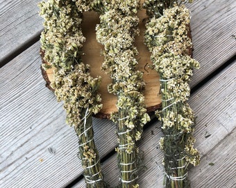 Può includere: Tre mazzi di fiori secchi, ciascuno legato con spago bianco. I fiori sono di colore beige chiaro e gli steli sono verdi. I mazzi sono disposti su una fetta di legno, su uno sfondo di legno stagionato.