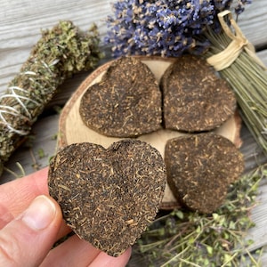 Puede incluir: Cuatro inciensos herbales prensados en forma de corazón, dos sobre una superficie de madera y dos en una mano. El incienso es de color marrón oscuro y tiene una textura rugosa.