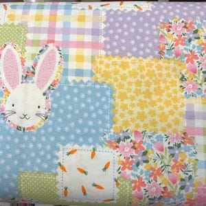 Peut inclure: Une courtepointe patchwork avec une applique de visage de lapin. La courtepointe présente des carrés de couleurs pastel et des motifs floraux, dont le rose, le jaune, le bleu et le vert. Le lapin a des oreilles roses et une bordure florale.