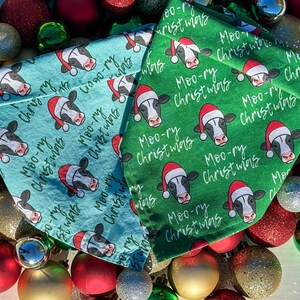 May include: Two Christmas-themed pet bandanas. One is teal, the other is green. Both feature a pattern of cow faces wearing Santa hats, with the text "Moo-ry Christmas" in a handwritten style. The bandanas are surrounded by Christmas ornaments.