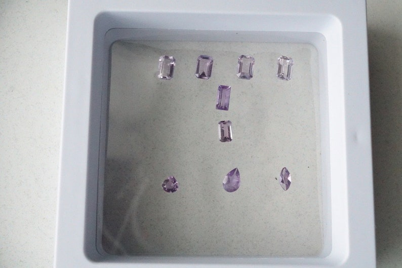 Puede incluir: Una colecci&oacute;n de ocho gemas de amatista en varias formas y tama&ntilde;os, expuestas en una bandeja blanca con un fondo gris claro.