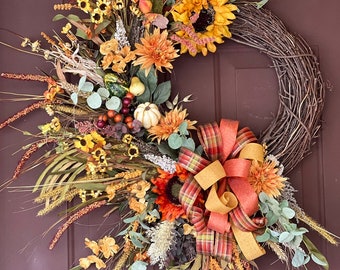 May include: A fall-themed wreath with a brown twig base, decorated with sunflowers, small yellow flowers, orange and yellow leaves, and a plaid and gold ribbon.