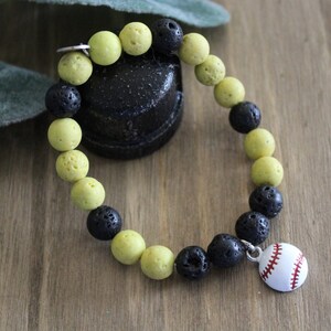 May include: A beaded stretch bracelet featuring alternating round yellow and porous black beads. The bracelet includes a small, round white charm with red baseball stitching details attached by a silver-toned metal jump ring.
