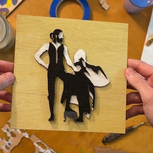 Puede incluir: Un recorte de madera de dos figuras, un hombre y una mujer, de pie sobre un fondo de madera claro. Las figuras son en blanco y negro, y el hombre está sosteniendo la mano de la mujer. La mujer lleva un vestido largo y el hombre lleva pantalones y un chaleco.