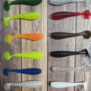 Puede incluir: Surtido de diez señuelos de pesca de plástico blando en varios colores, incluyendo verde, amarillo, naranja, azul, negro y blanco. Los señuelos están dispuestos en dos filas de cinco sobre un fondo de madera.