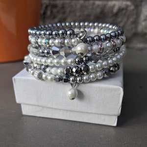 May include: A stack of seven bracelets featuring white pearls, silver beads, and dark grey beads. The bracelets are arranged in a circle and are resting on a white box.