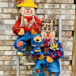 May include: Two scarecrows dressed in denim overalls and plaid shirts are sitting on a wooden ladder. The scarecrows have straw hair and painted faces. The scarecrow on the top of the ladder has a yellow hat and the scarecrow on the bottom has a brown and white plaid hat. The ladder is leaning against a brick wall.