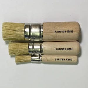 May include: Three paint brushes with natural bristles and wooden handles. The handles are labelled with the text "18 BRITISH MADE", "12 BRITISH MADE", and "6 BRITISH MADE". The brushes are arranged in a row, with the largest brush on top.