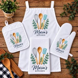 May include: A white kitchen set including an apron, a square potholder, a waffle-weave towel, and an oven mitt. Each piece features a graphic of a whisk, wooden spoon, and blue spatula with green leaves and the text Maria's KITCHEN.