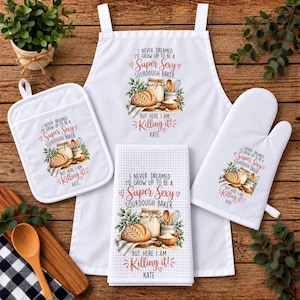 May include: A white kitchen set including an apron, a square pot holder, and a rectangular waffle-weave towel. Each piece features a graphic of a sourdough loaf, a jar, and utensils with the text: I NEVER DREAMED I'D GROW UP TO BE A SUPER SEXY SOURDOUGH BAKER BUT HERE I AM KILLING IT! KATE.