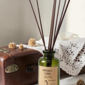 May include: A green glass reed diffuser bottle with brown reeds. The label reads "Midday calm" with text below. The bottle is placed near a brown vintage suitcase and dried flowers. The diffuser is 4 oz / 113g.