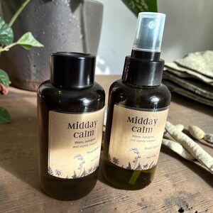 May include: Two dark brown glass bottles with black caps. One is a spray bottle. Both have labels that read "Midday Calm" with text below. The bottles are filled with a green liquid. The labels also say "Warm, indulgent, and slightly tropical."