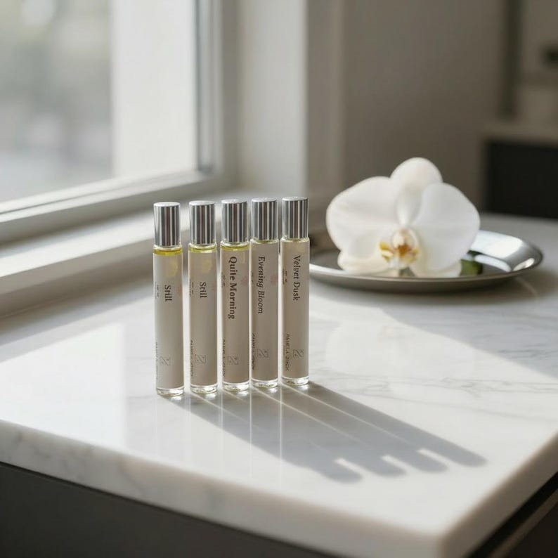 May include: Five perfume bottles with silver caps and labels that read "Still", "Still", "Quite Morning", "Evening Bloom", and "Velvet Dusk". A white orchid sits on a silver dish. The bottles are on a white marble surface.