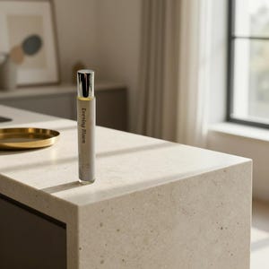May include: A slender perfume bottle with a silver cap and a light gray label. The label reads "Evening Bloom" in a vertical orientation. The bottle is standing upright on a light-colored countertop, with a gold-colored dish in the background.